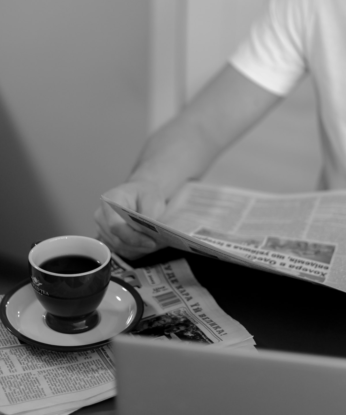Person liest Zeitung am Tisch mit Laptop und Tee.